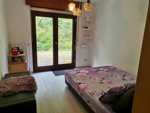 Ein Schlafzimmer mit einem Bett, einem kleinen Tisch, einem Stuhl, einem Heizkörper und Blick ins Grüne durch die Tür.
