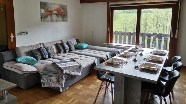 Een moderne woonkamer met een modulaire bank, een eettafel voor vier personen, en een groot raam met uitzicht op de omgeving.