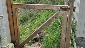 Een houten poort met een metalen grendel, gelegen in een tuin met een witte hekken en begroeiing op de achtergrond.