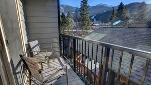 A balcony with a wooden bench and a metal railing, overlooking a mountainous landscape with trees and a house. - Creekside Mountain View Studio!Patio + BBQ,Pool,Parking & Walk to Shops and Lakes! (Whistler)