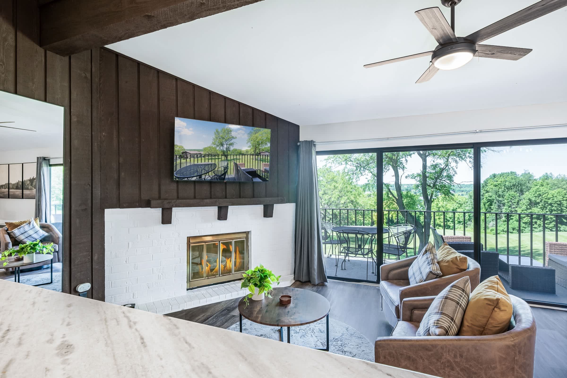 Un salon moderne doté d’une cheminée, d’un ventilateur de plafond et d’une grande fenêtre donnant sur un jardin.
