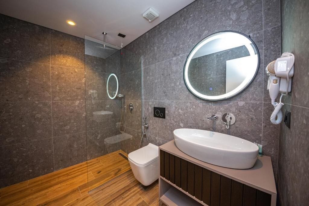 A modern bathroom with a wooden floor, a white sink, a mirror, a handheld hair dryer, and a glass shower enclosure.