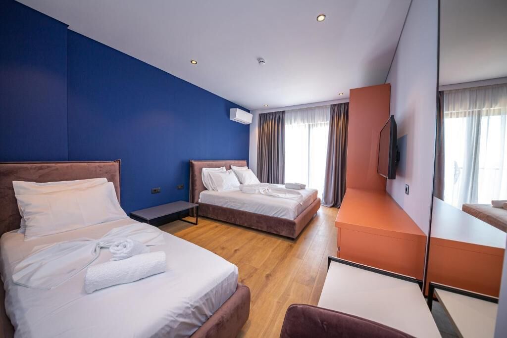 A hotel room with two beds, a desk, a TV, and a large window with curtains.
