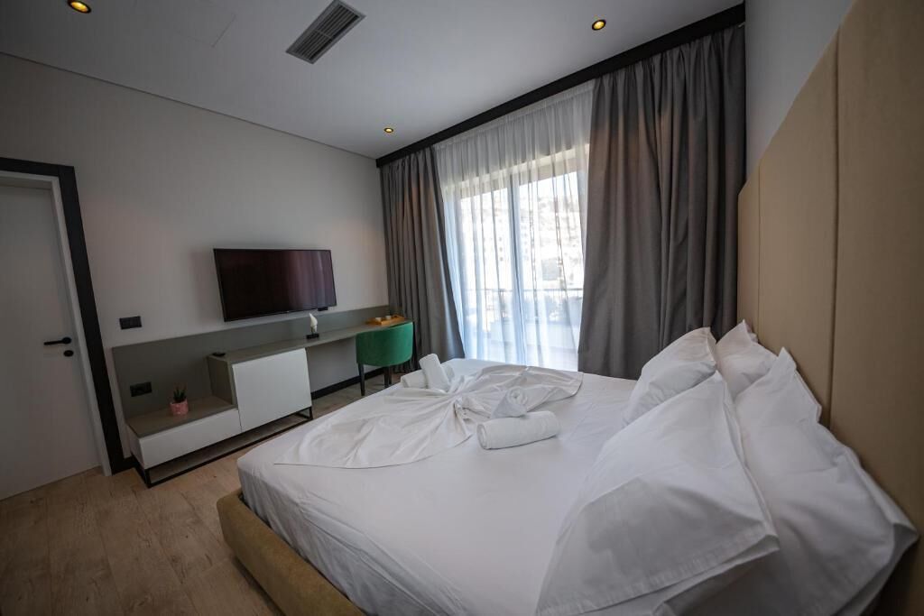 A modern hotel room with a large bed, a flat-screen TV mounted on the wall, a desk with a chair, and a window with curtains.
