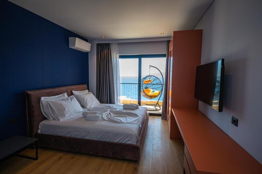 A modern hotel room with a large bed, a TV, and a balcony with a view of the sea.