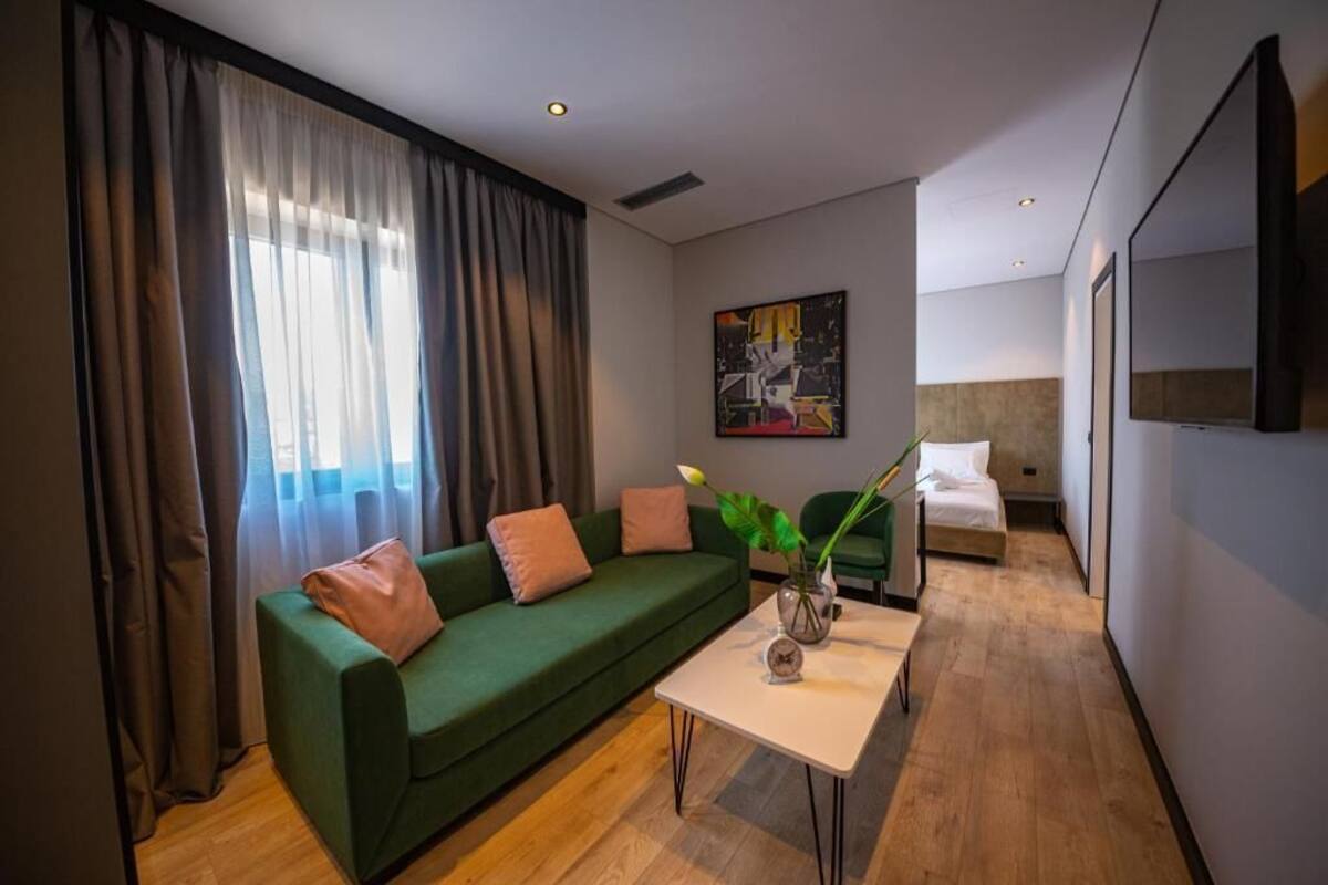A modern hotel room with a green sofa, a small table with a vase, a bed with white bedding, and a large painting on the wall.