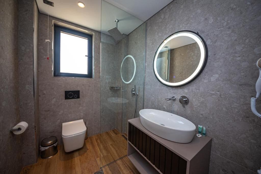 A modern bathroom with a glass-enclosed shower, a large round mirror, and a wooden vanity.