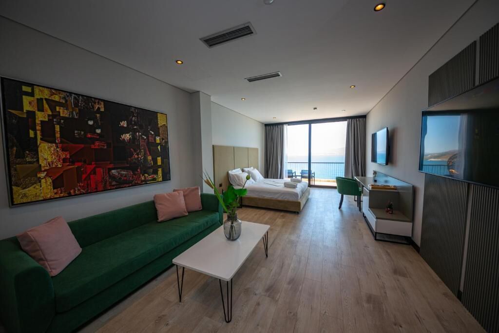 A modern hotel room with a green sofa, a white coffee table, a large abstract painting, a bed, a TV, and a view of the outdoors.