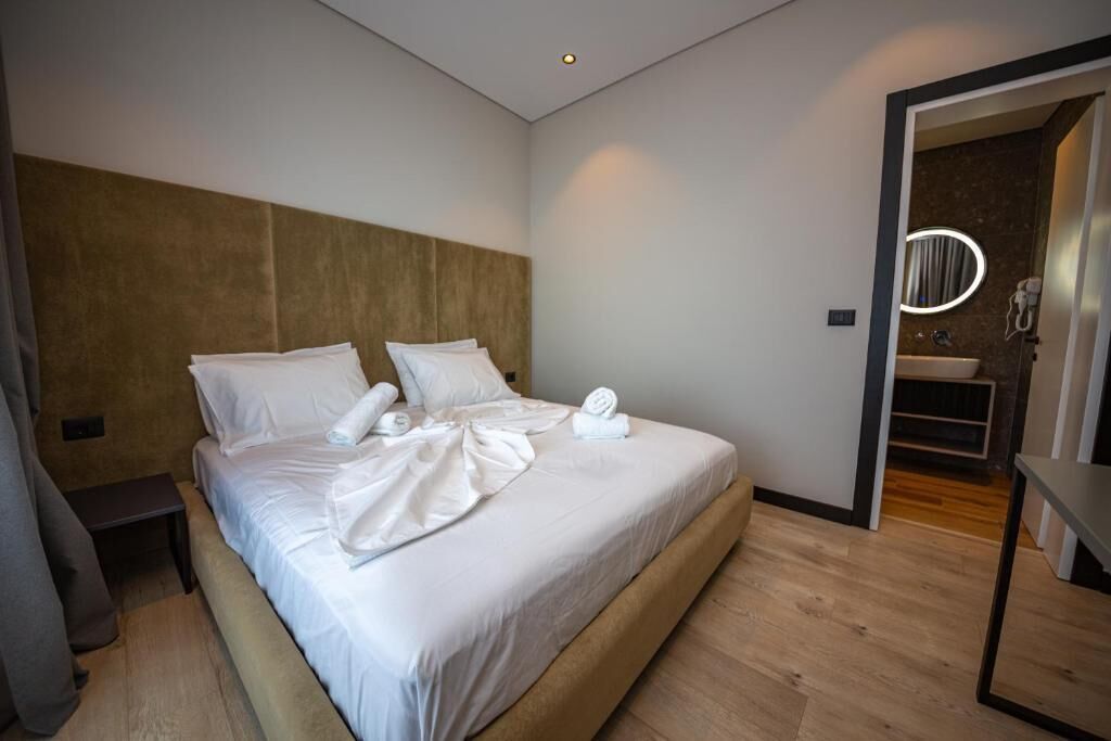 A modern hotel room with a large bed, a wooden headboard, a bedside table, and a bathroom visible through an open door.