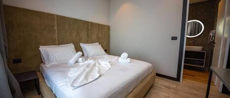 A modern hotel room with a large bed, a wooden headboard, a bedside table, and a bathroom visible through an open door.