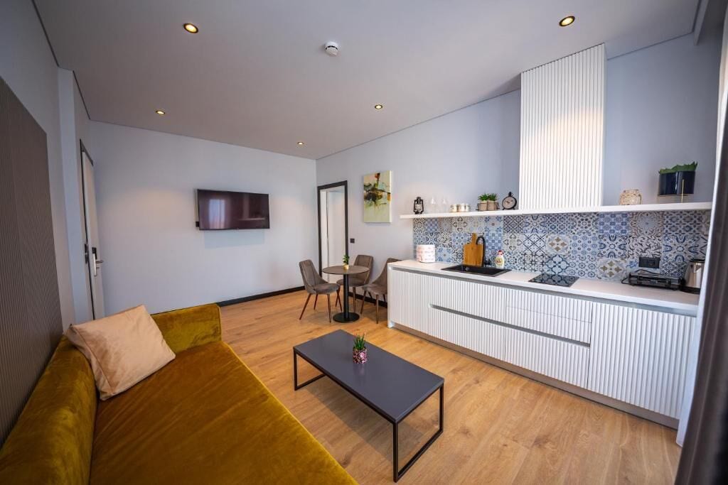 A modern living room with a sofa, a coffee table, and a kitchen area with a tiled backsplash and a white cabinet.
