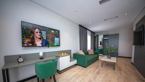 A modern living room with a flat-screen TV mounted on the wall, a green sofa, a white coffee table, and a small desk with a chair. - Apart-Hotel Lili2 (Sarandë)