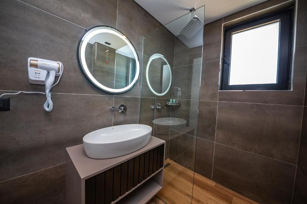 A modern bathroom with a large round mirror, a white sink, a handheld hair dryer, and a shower area with a glass door.