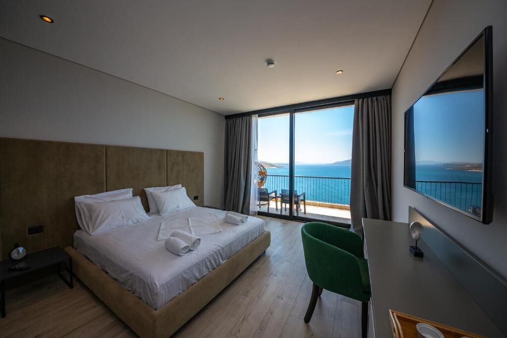 A modern hotel room with a large bed, a desk, and a balcony with a view of the sea.