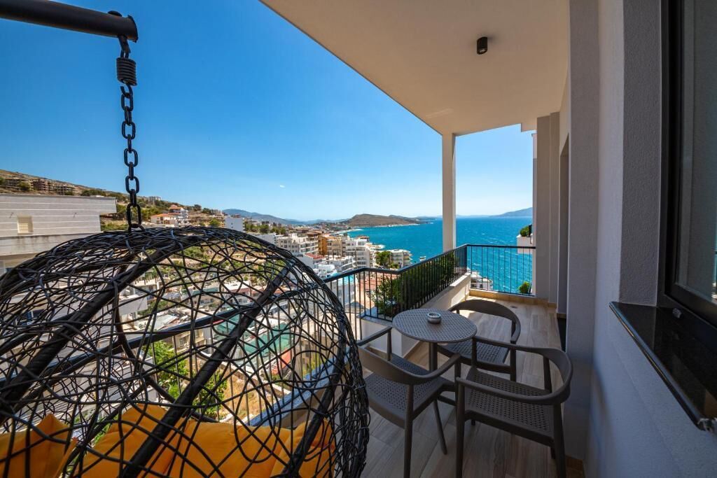 A balcony with a hanging chair, a small table, and a view of the sea and buildings.