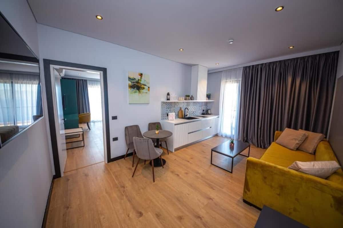 A modern living room with a yellow sofa, a dining area with chairs, a kitchen with a white countertop, and a view through an open doorway.