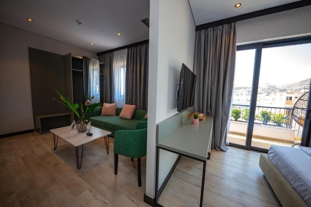 A modern hotel room with a green sofa, a small table with a vase, a TV on the wall, and a balcony with a view of the city.