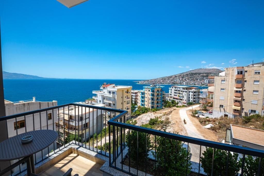 A balcony with a table and chairs, overlooking a coastal town with buildings and a clear blue sky.