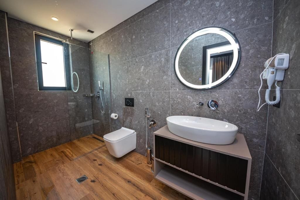 A modern bathroom with a wooden floor, a white sink, a round mirror, and a handheld showerhead.