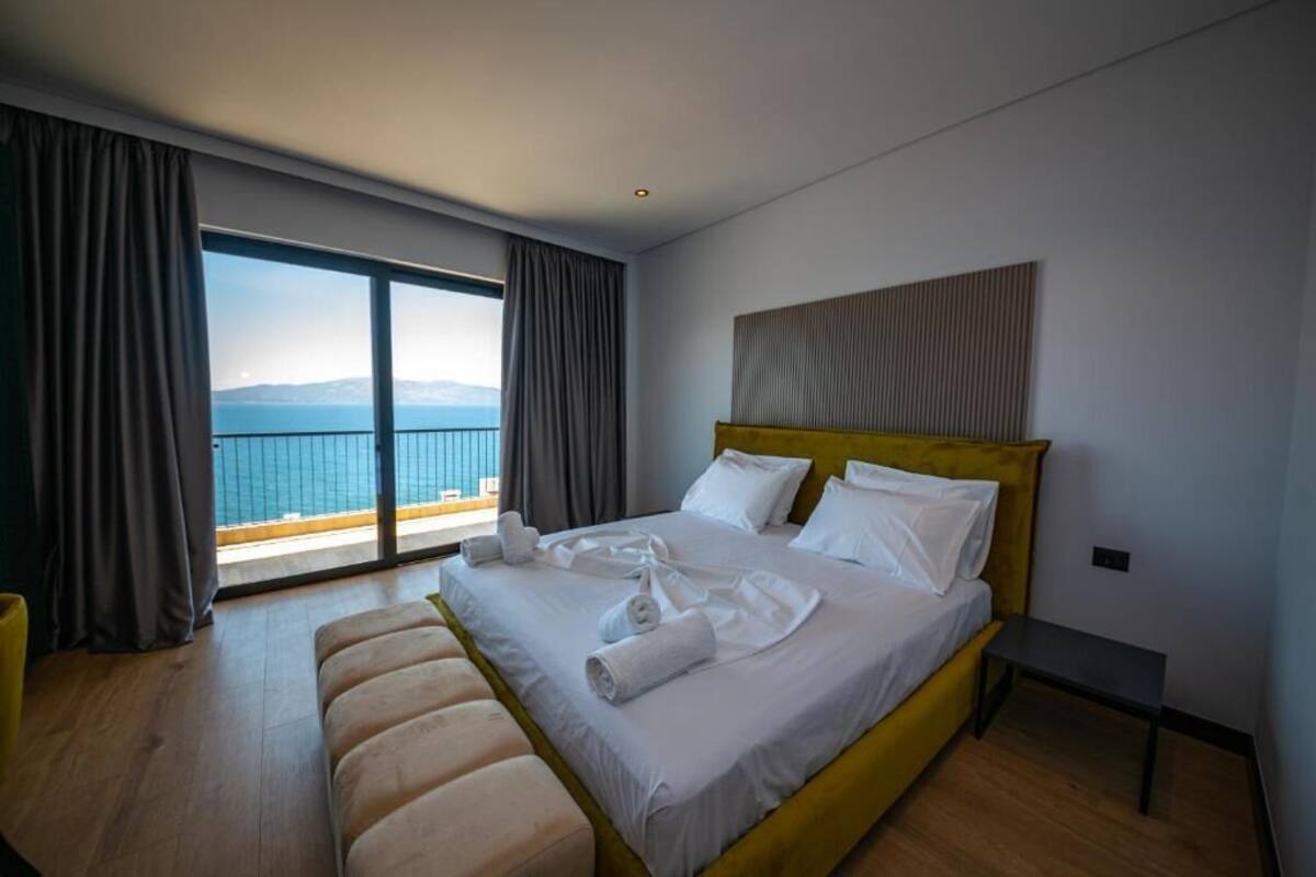 A modern hotel room with a large bed, a small bedside table, and a view of the sea through a sliding glass door.