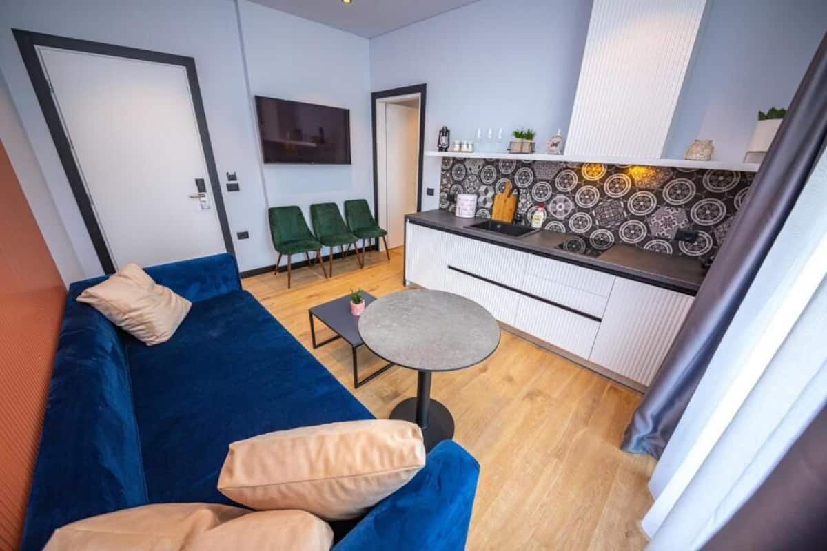 A compact living space with a blue sofa, a small round table, and a kitchenette area with patterned backsplash and white cabinets.