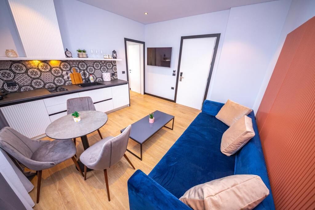 A modern living space with a blue sofa, grey chairs, a small round table, and a kitchen area with patterned backsplash and a countertop.