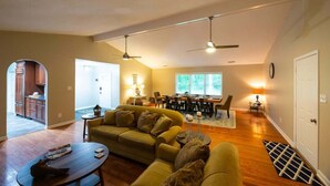 A spacious living area with a sofa, coffee table, and dining table. There are ceiling fans, a clock, and a door visible. - Lake T Retreat - Sunset - Pool - Putting Green! (Northport)