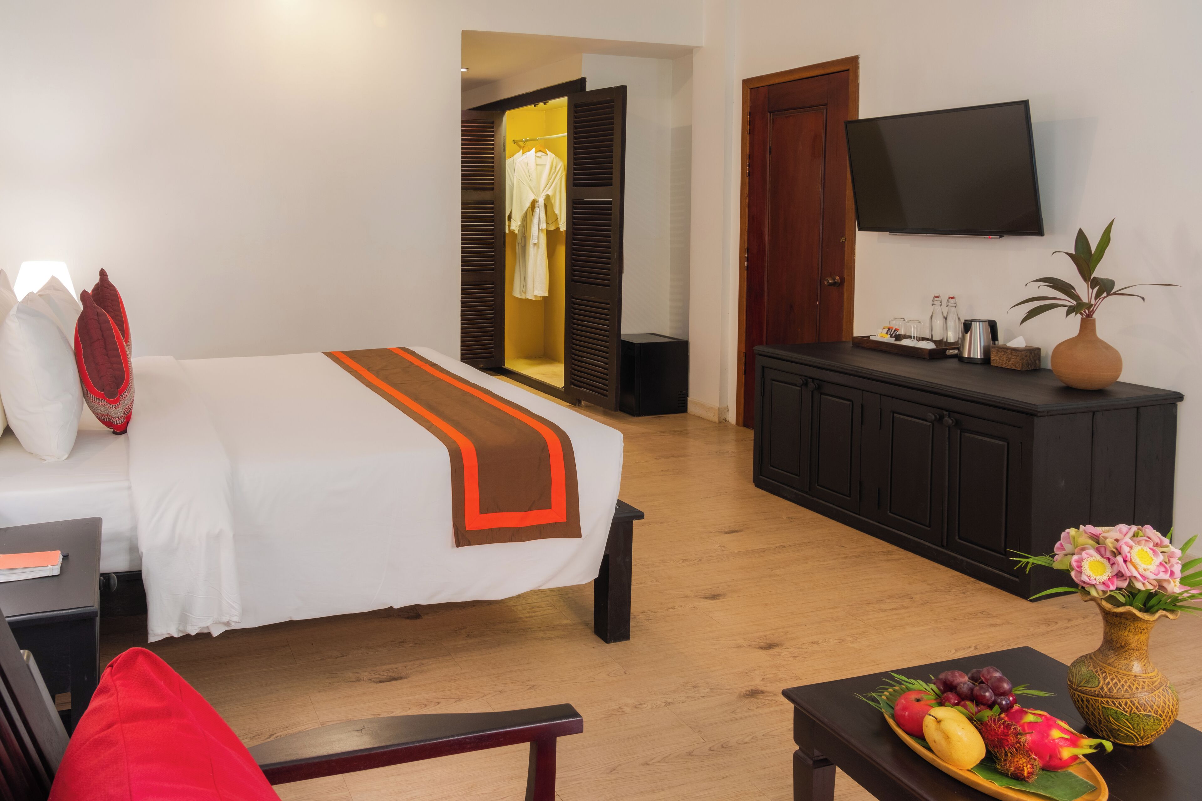 A hotel room with a bed, a desk, a TV, a wardrobe, and a dining table with fruit.