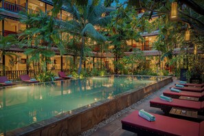 Zona de piscina con sillones de descanso y piscina reflectante, rodeada de plantas tropicales y un edificio de varios pisos con balcones.