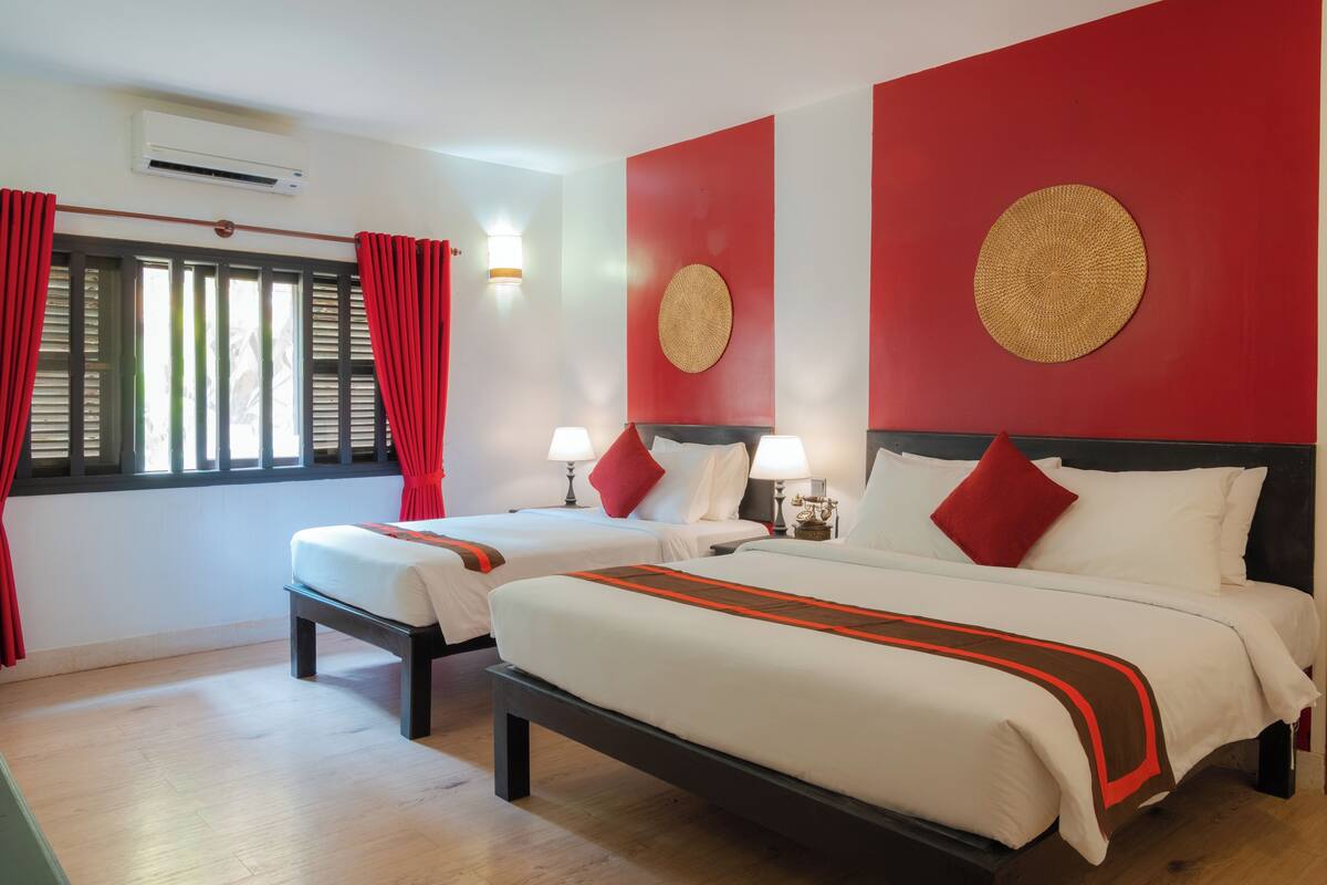Una camera d'albergo con due letti, tende rosse, un condizionatore d'aria a muro e un ornamento circolare sulla parete.