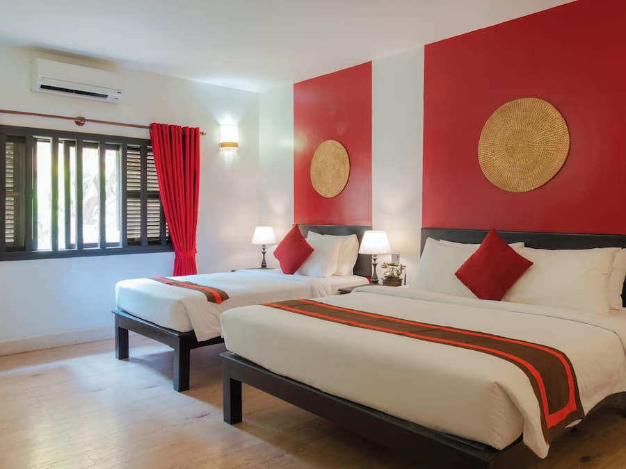 Habitación de hotel con dos camas, cortinas rojas, aire acondicionado de pared y un adorno redondo en la pared.