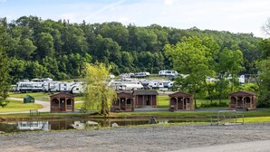 Um parque de campismo com veículos recreativos, pequenas cabanas de madeira, um lago e uma colina arborizada ao fundo.