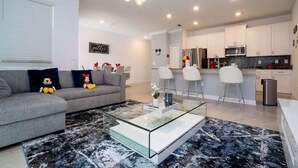 Un saló modern amb un sofà gris, una taula de centre de vidre i una zona de cuina amb armaris blancs i un rentapeus d'acer inoxidable.