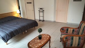 Une chambre à coucher comprenant un lit, une petite table avec une plante, deux fauteuils rembourrés, un porte-vêtements suspendu et une porte.