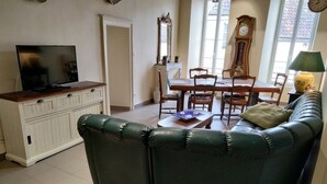 Un salon avec un canapé en cuir vert, une table basse en bois, un téléviseur posé sur un meuble, une table à manger avec des chaises et une grande fenêtre donnant sur des immeubles.