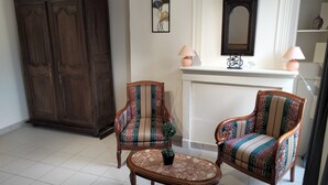 Un salon comprenant deux fauteuils rembourrés, une table basse en bois, un grand meuble en bois foncé, une cheminée agrémentée de deux lampes murales et une œuvre d’art encadrée au-dessus de la cheminée.