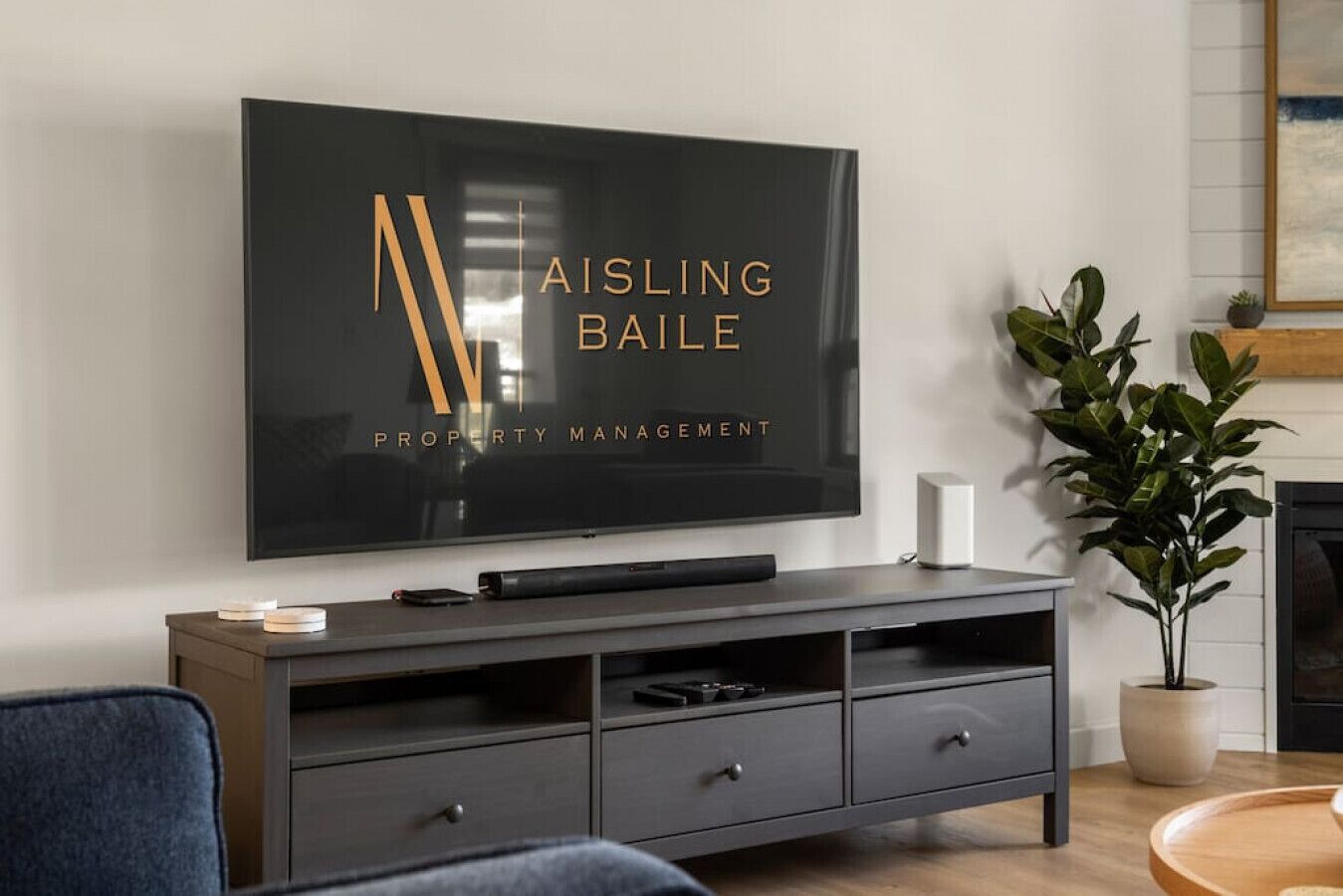 A modern living room with a flat-screen TV displaying a logo, a dark grey entertainment unit, a blue chair, and a potted plant.