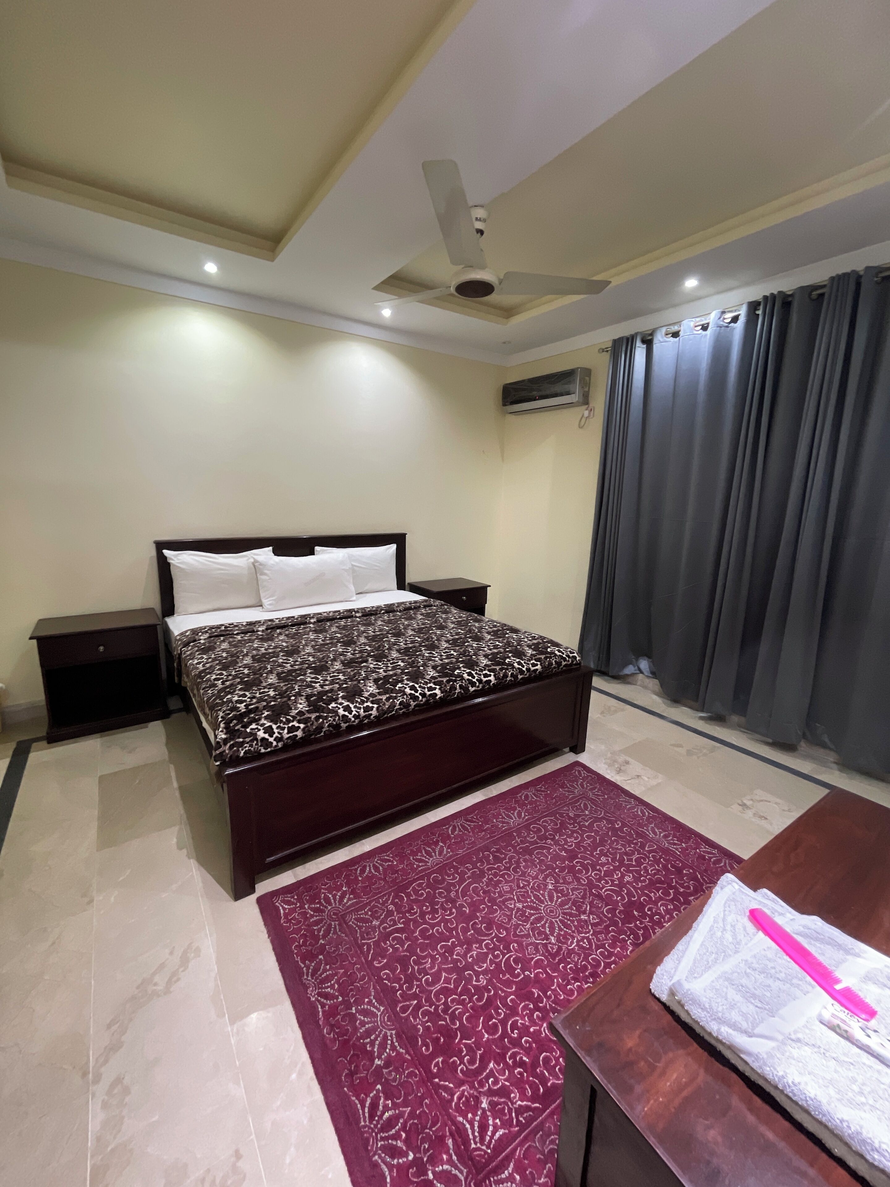 A bedroom with a bed, nightstands, a ceiling fan, an air conditioning unit, a window with curtains, and a patterned rug.