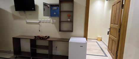 Quarto com televisão de tela plana presa na parede, um refrigerador branco pequeno, uma porta de madeira e um móvel azul com uma planta em vaso.