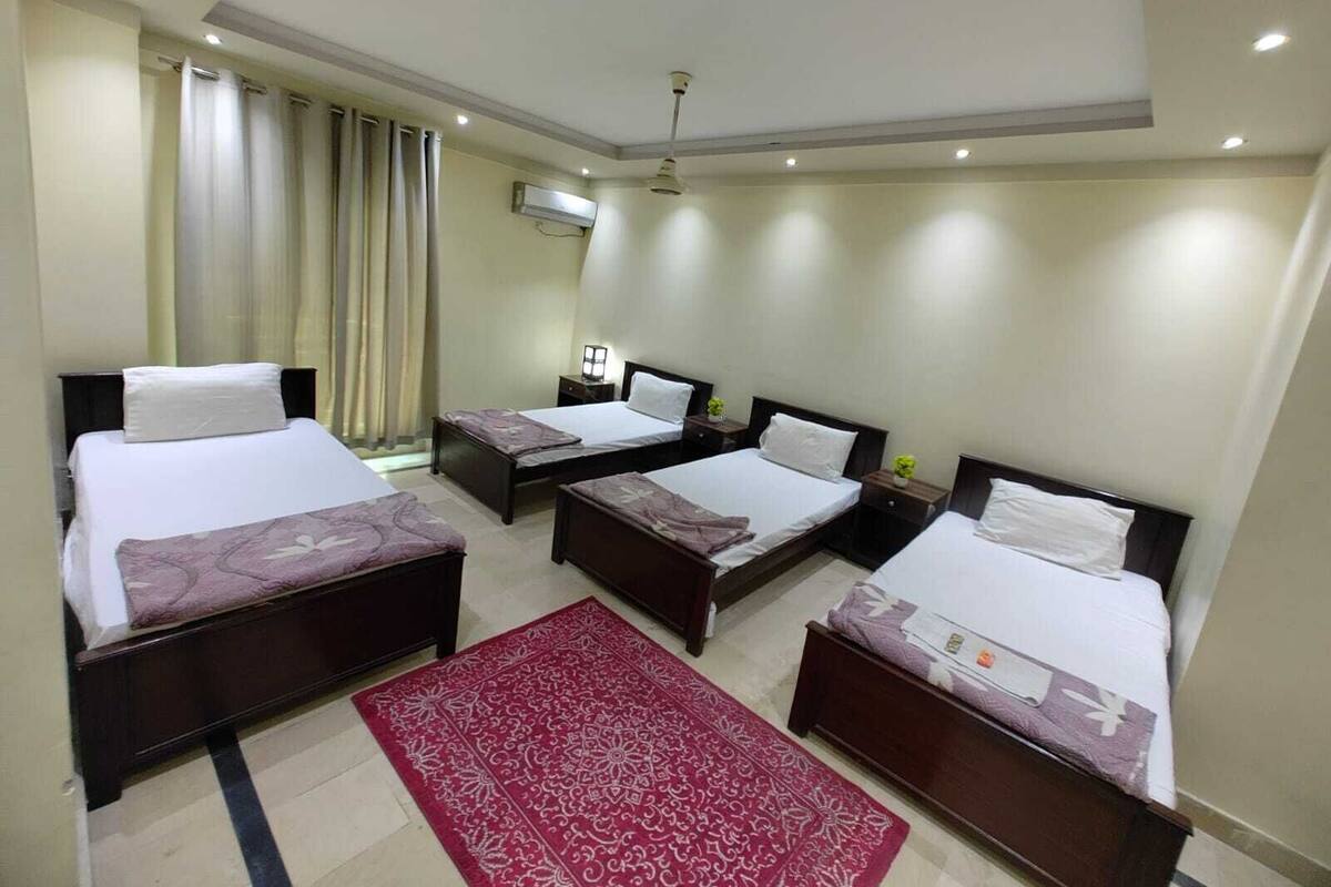 Ein Hotelzimmer mit drei Betten, einem roten Teppich mit Muster, einem Deckenventilator und einer Klimaanlage.