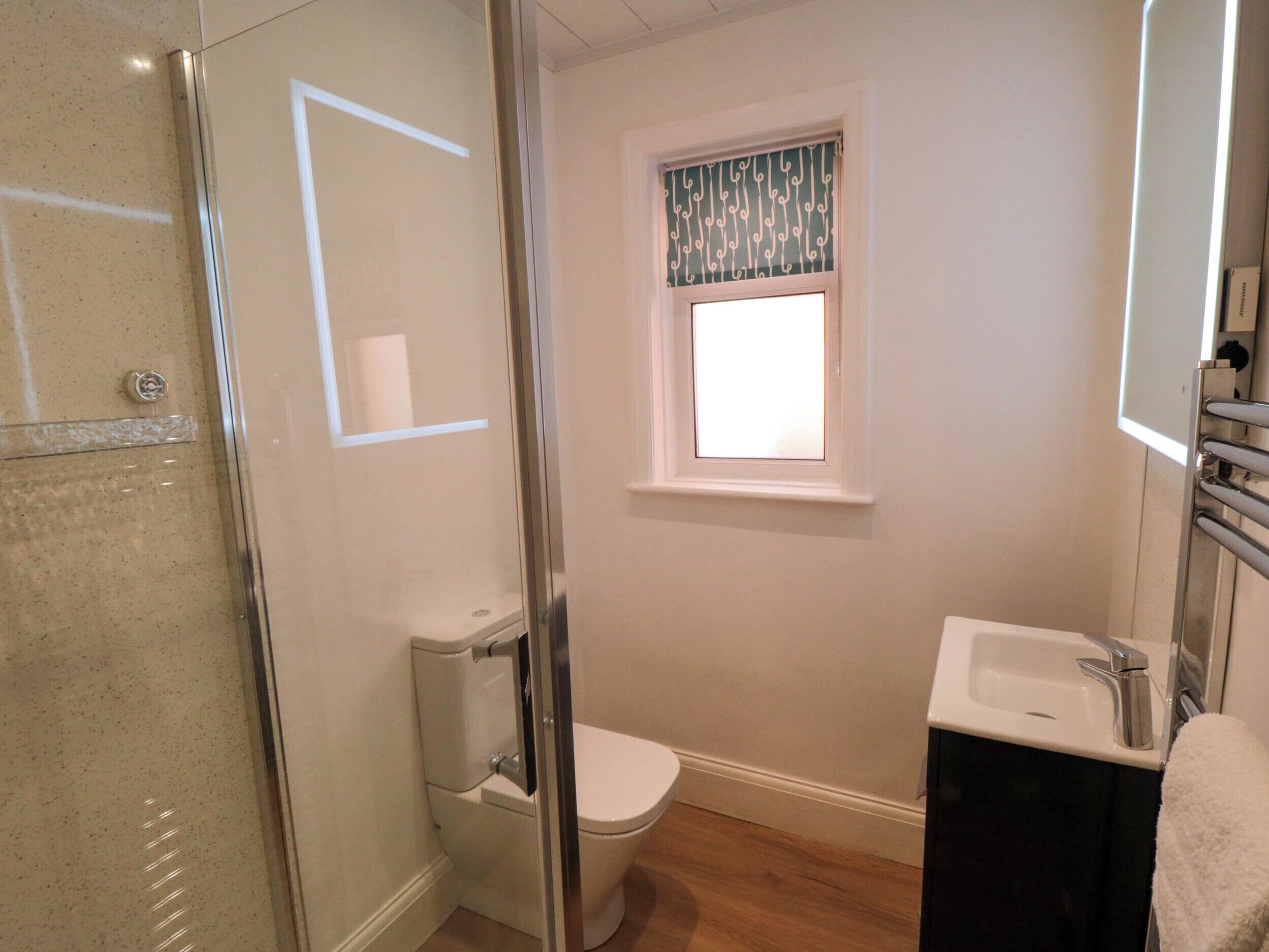 A bathroom with a glass shower enclosure, a toilet, a window with a patterned curtain, and a sink with a towel.