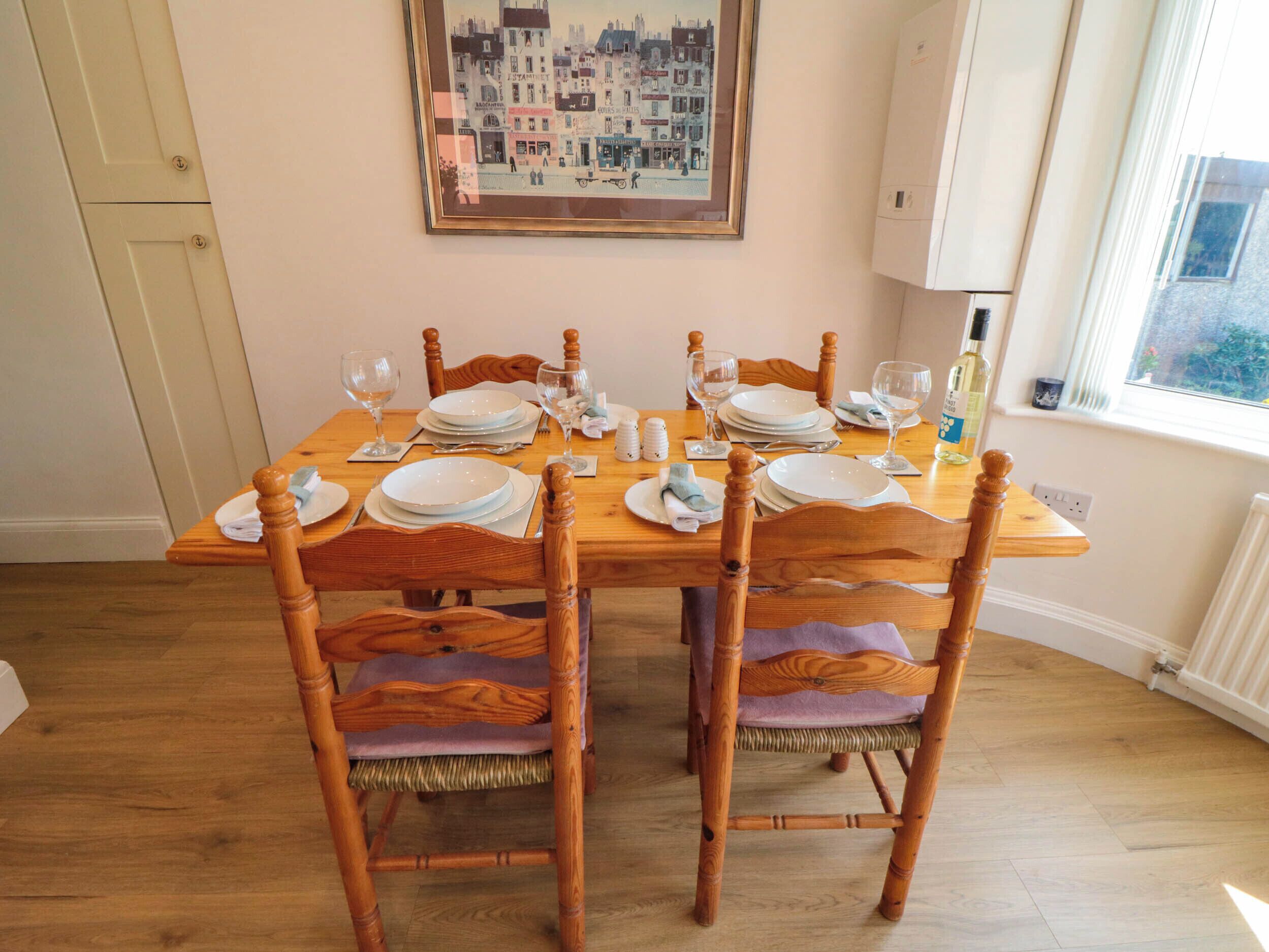 A dining table set for four with wooden chairs, a framed cityscape picture on the wall, and a window with a view.