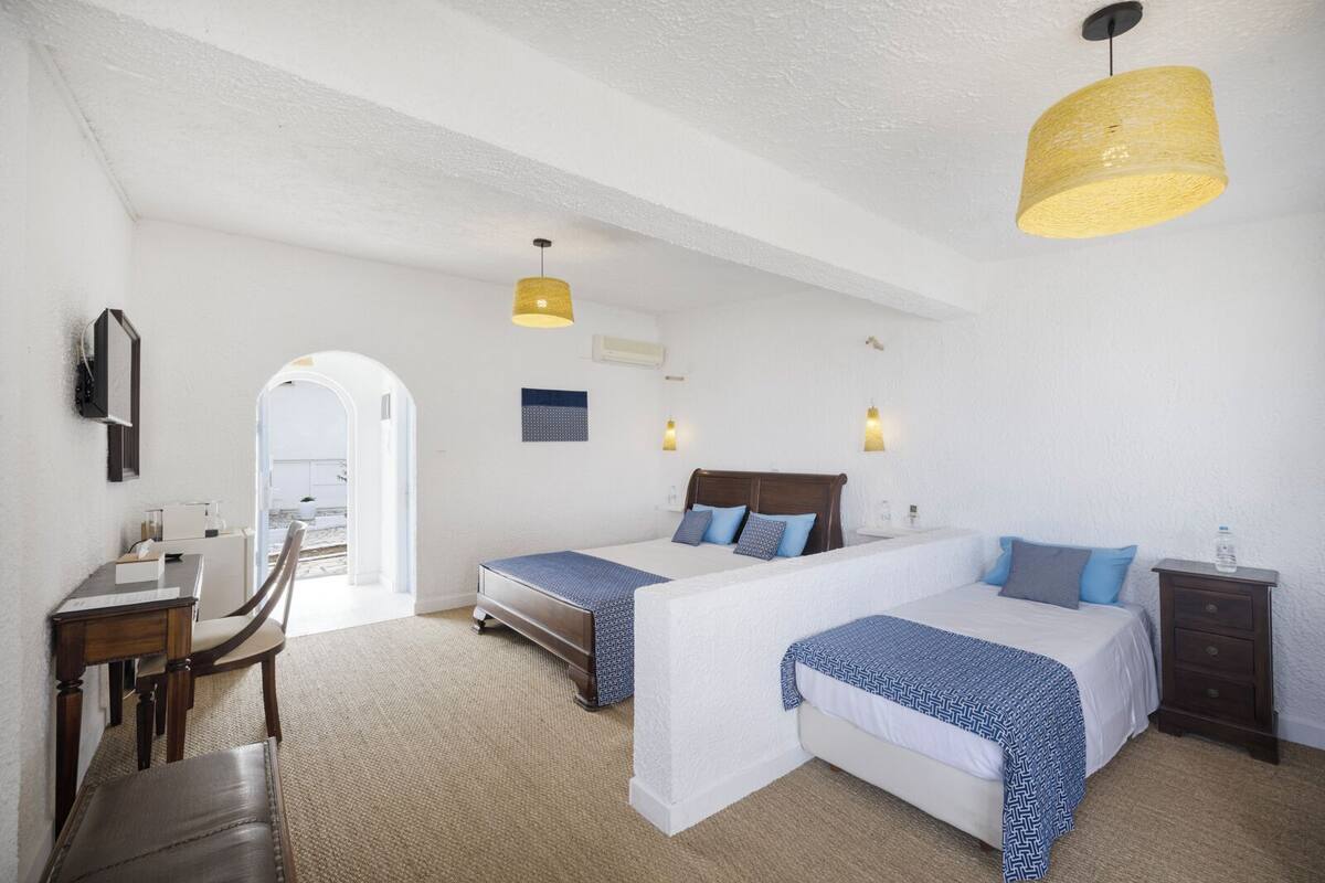 A hotel room with two beds, a desk, and a chair. There is a large window and a door leading outside.
