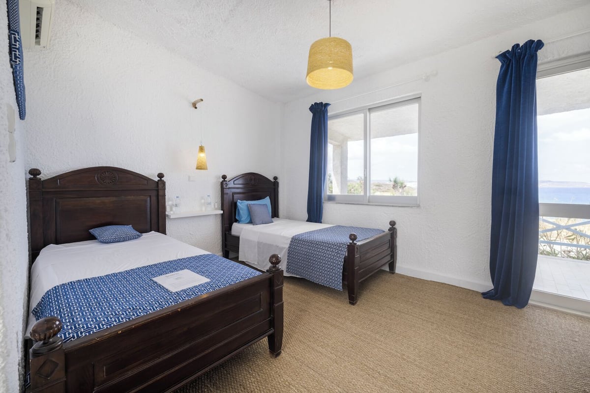 A room with two wooden beds, a hanging light fixture, a window with curtains, and a view of the sea.