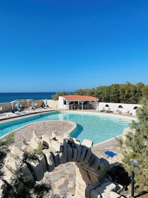 A pool area with a stone structure, lounge chairs, and a building with a red roof. - Lassion Hotel (Sitia)