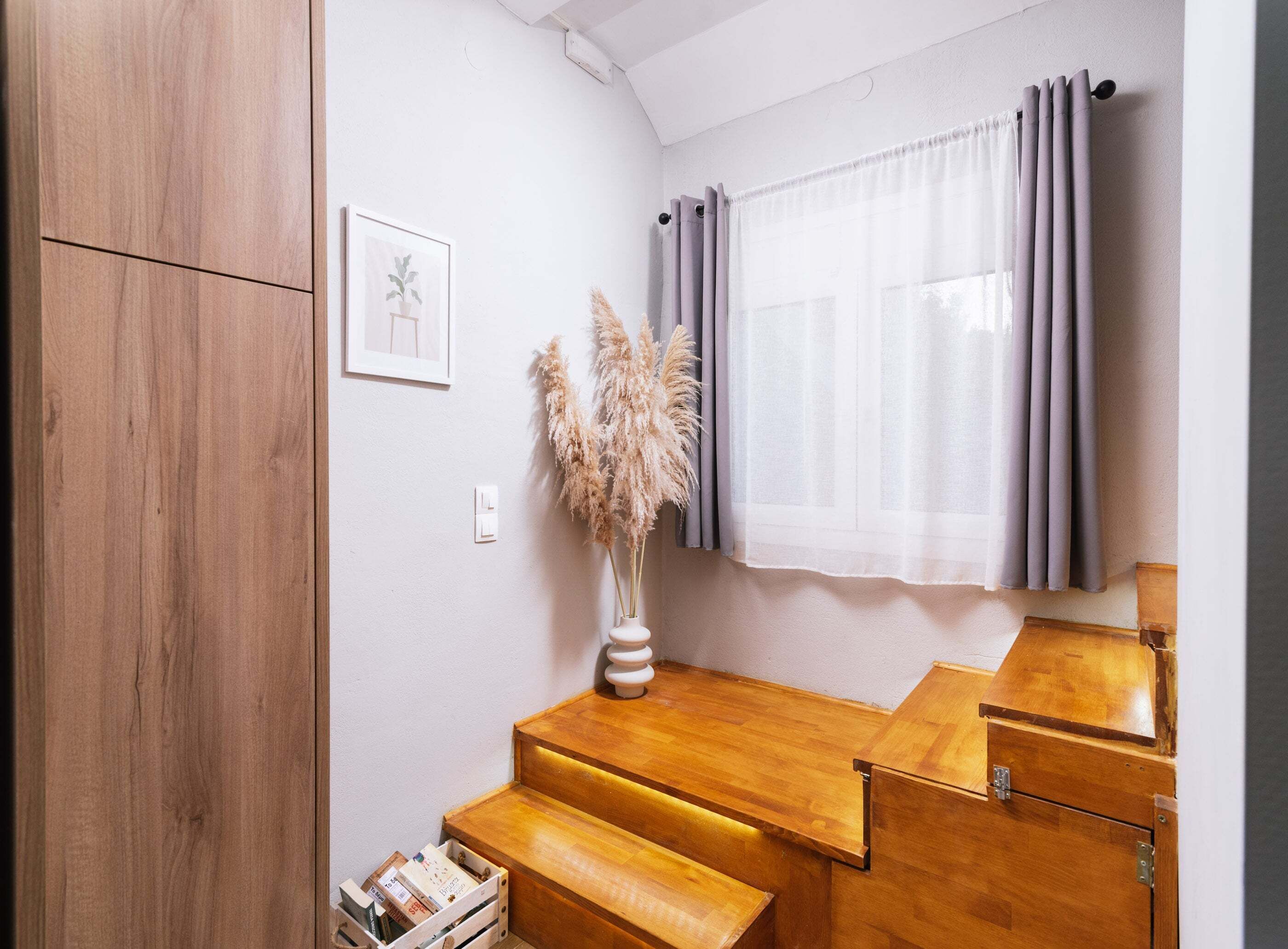 Una habitación compacta con un armario de madera, un pequeño escritorio de madera con cajones, un cuadro enmarcado en la pared y una ventana con cortinas.
