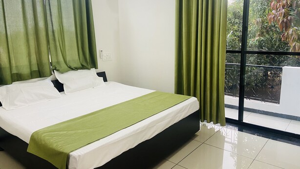 A hotel room with a bed, green curtains, a window with a view of greenery, and an air conditioning unit.