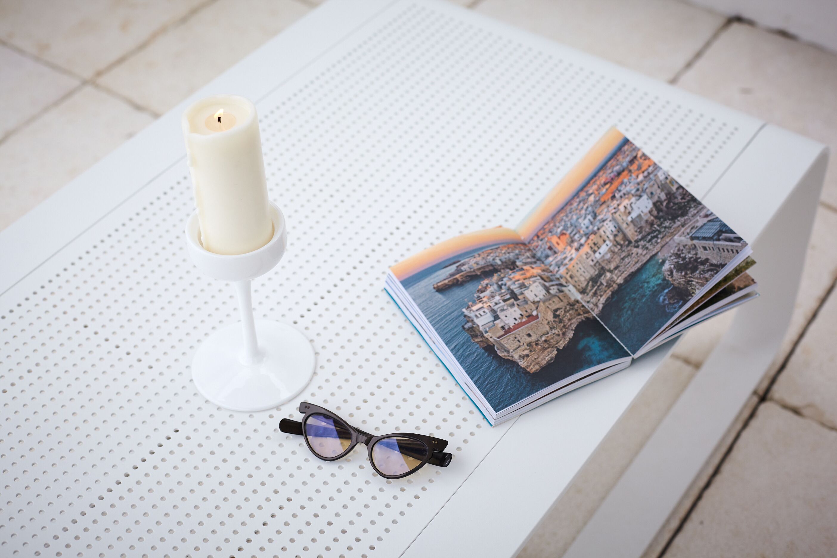 A white table with a candle, glasses, and an open magazine featuring a coastal cityscape.