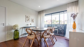 A modern dining area with a round table and six chairs, a green vase with white flowers, and a view of buildings through large windows. - Nomads Lux - 2BDR Parking Flores Porto (Porto)