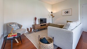A modern living room with a grey sofa, a wooden TV stand, a small table with a book, and a woven basket. - Nomads Lux - 2BDR Parking Flores Porto (Porto)